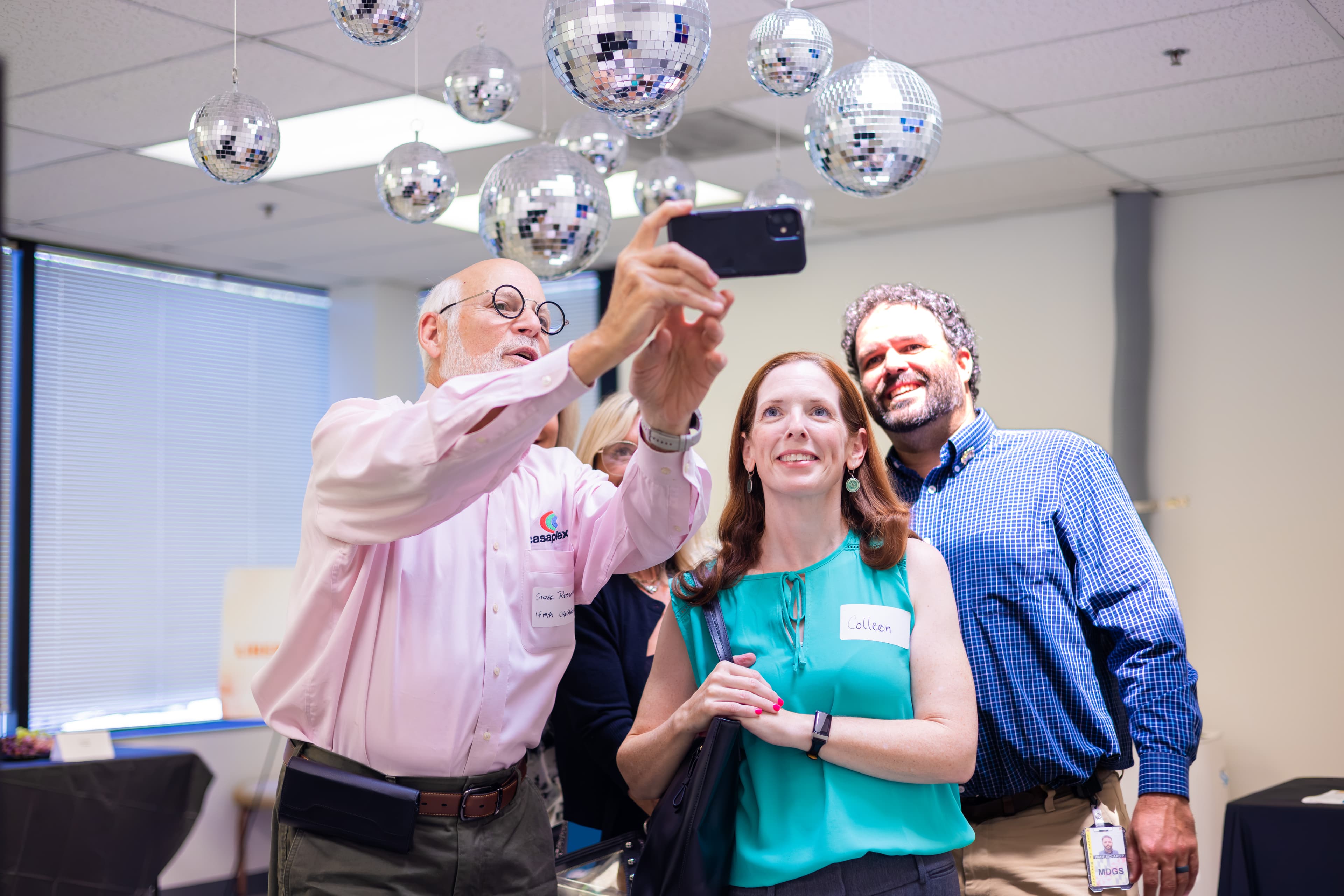 A group of colleagues smiling for a selfie under hanging disco balls in an office.