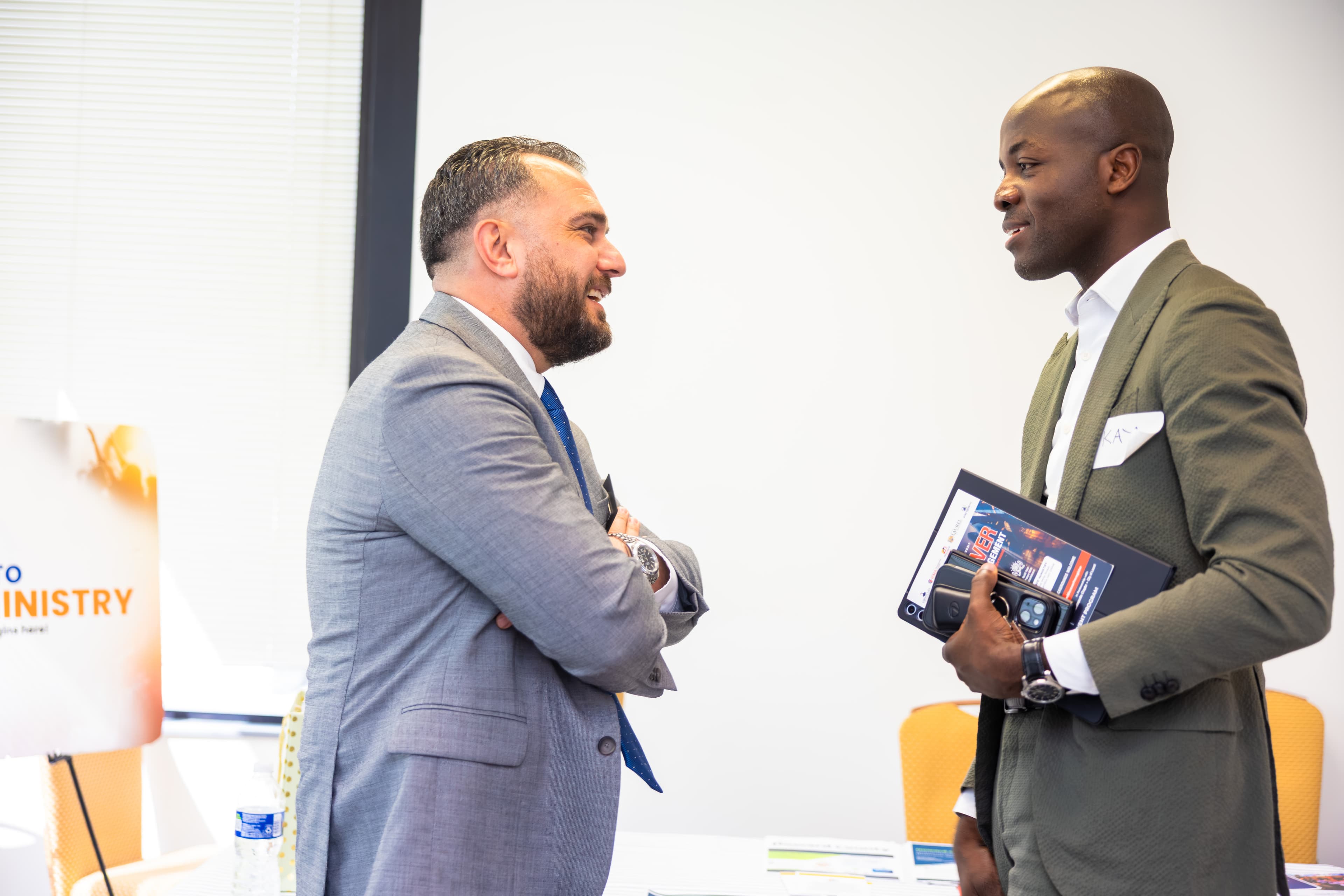 Two men in business suits, one grey and one olive, smiling during a professional conversation.
